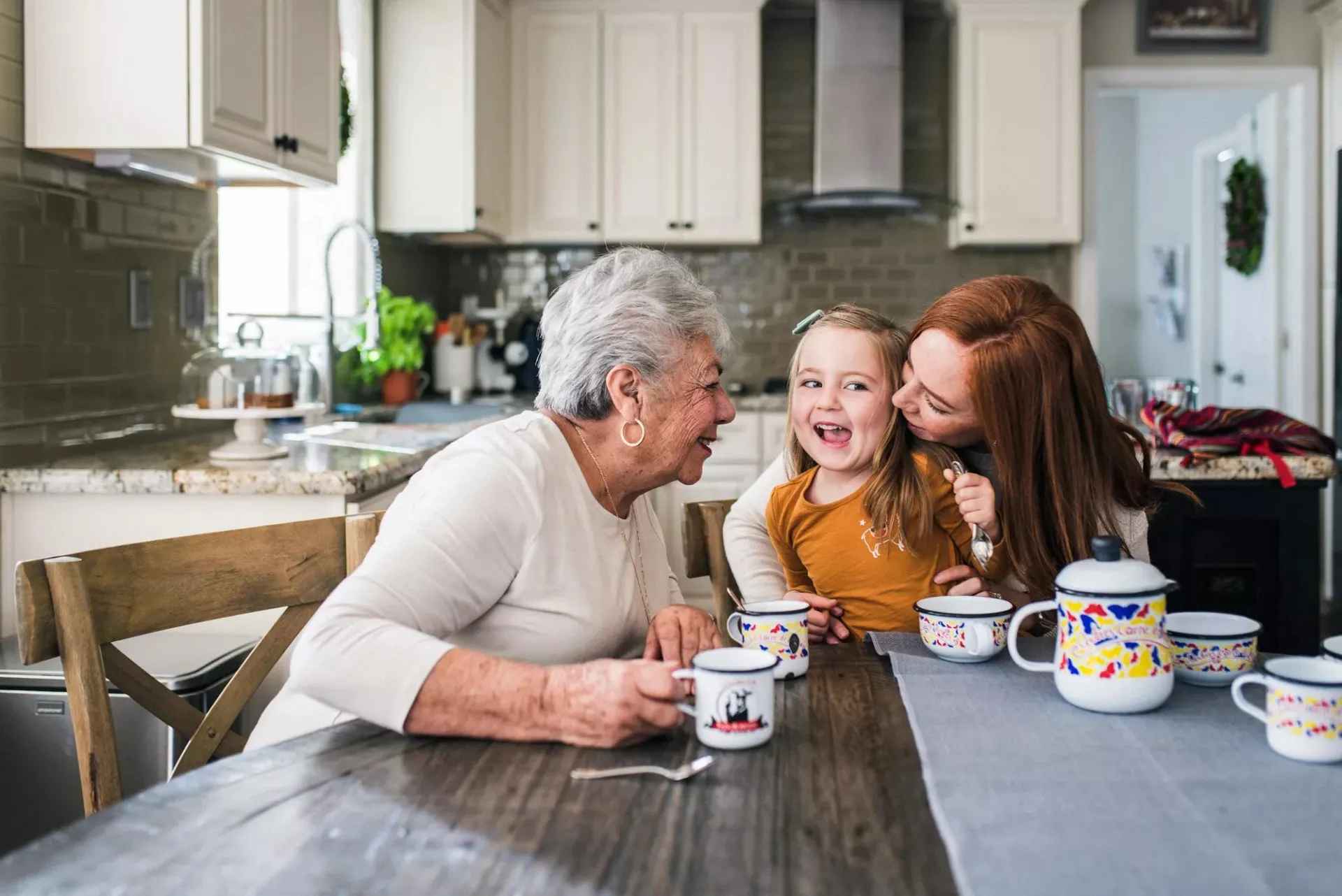 Mother kissing daughter on the cheek at kitchen table with grandma laughing.